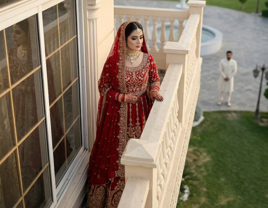 Bride in red wedding dress standing on balcony at luxury photoshoot venue near Islamabad with scenic garden view